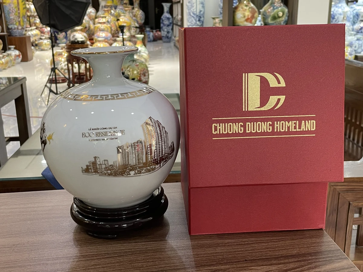 Bình tròn trắng in hình công trình trên đế nâu cạnh hộp đỏ CHUONG DUONG HOMELAND