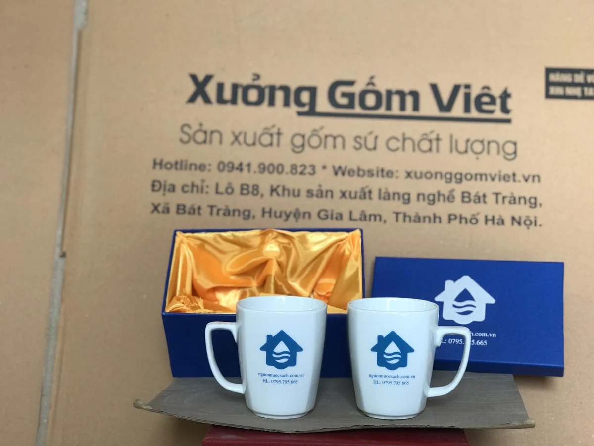 Hai ly sứ quai vuông in biểu tượng xanh cạnh hộp hai ly lót vàng