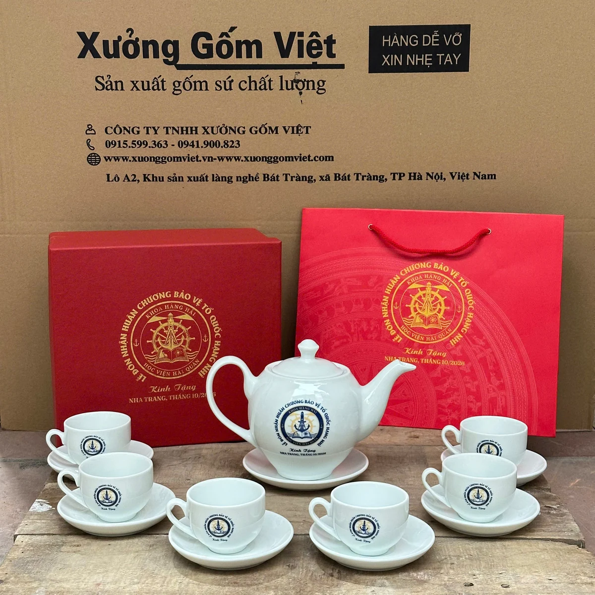 Bộ trà trắng in biểu trưng xanh vàng, đặt cùng hộp và túi đỏ trong ảnh đơn hàng