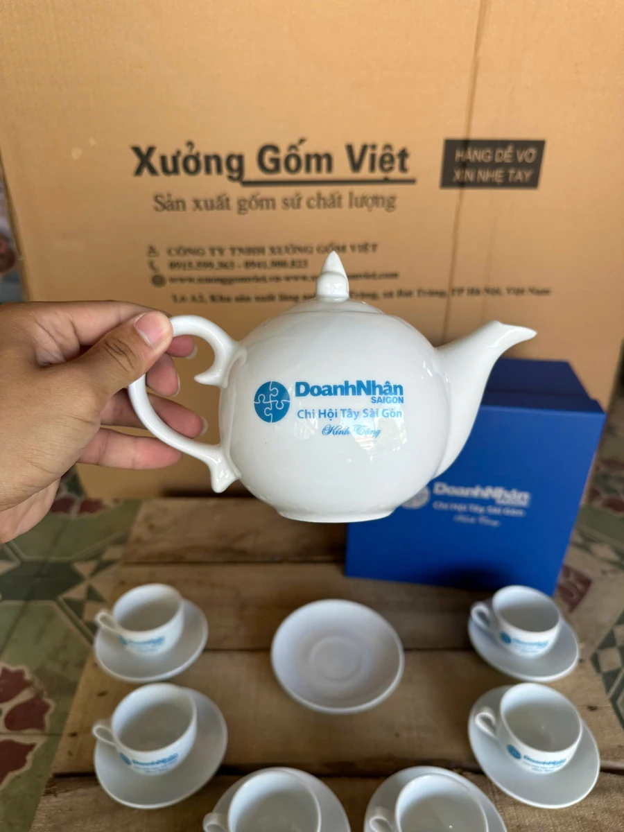 Tay cầm ấm sứ trắng có nhận diện Doanh Nhân Sài Gòn cạnh hộp xanh