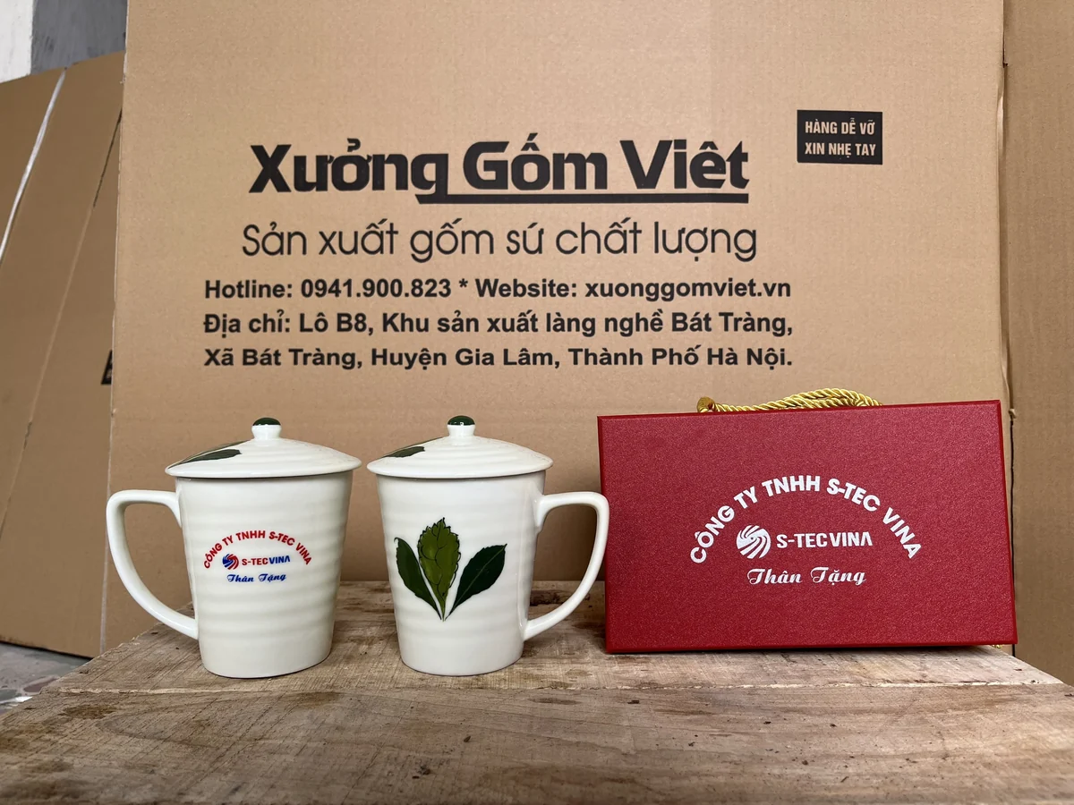 Hai ly sứ có nắp núm xanh, in nhận diện S-TEC VINA và hoa lá cạnh hộp đỏ