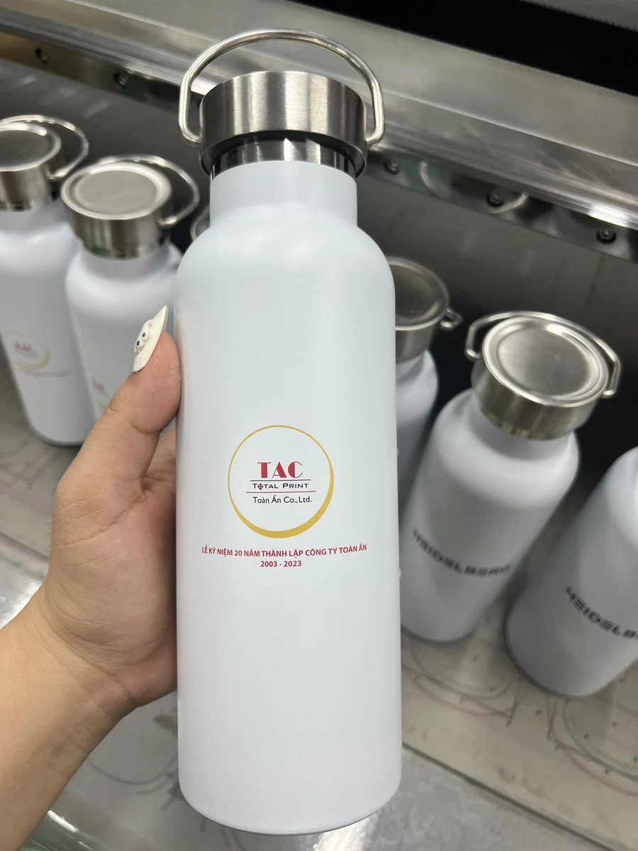 Bình trắng có nắp kim loại, quai gập và logo TAC được cầm trên tay