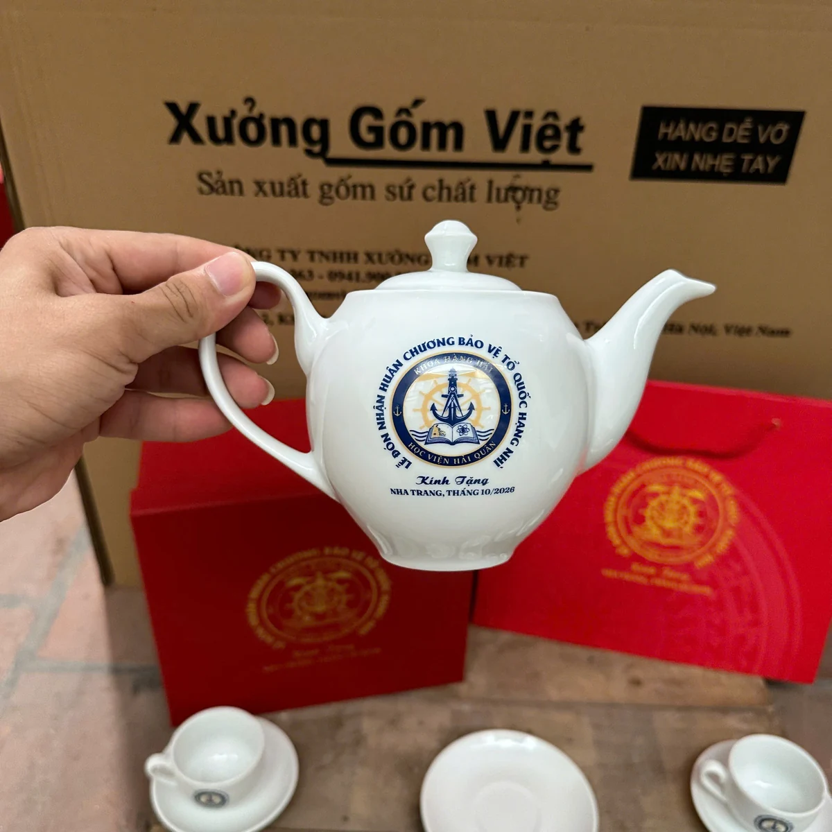 Một người cầm quai ấm trắng có nắp, vòi cong và biểu trưng in phía trước