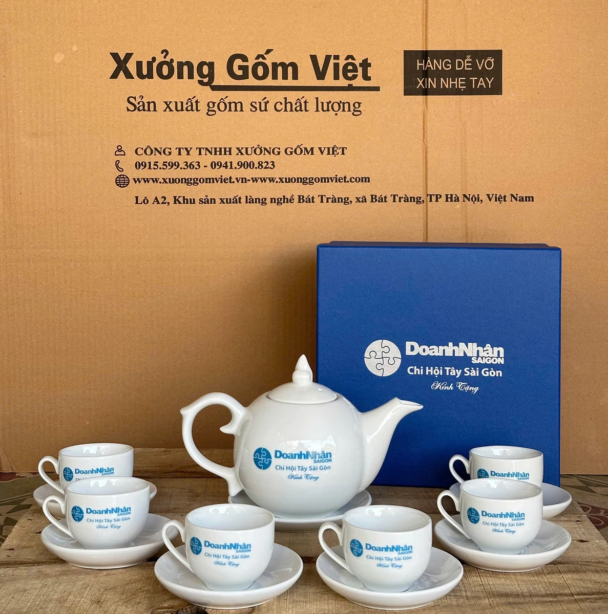 Một ấm sứ trắng có nắp cùng sáu chén và đĩa trước hộp xanh in Doanh Nhân Sài Gòn