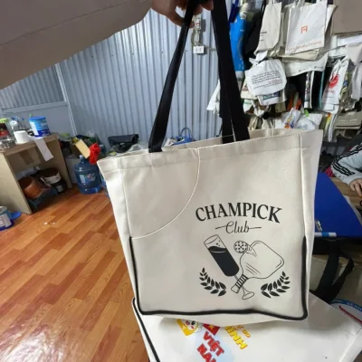 Tư liệu cầm hai quai đen của túi canvas kem với hình CHAMPICK Club ở mặt trước