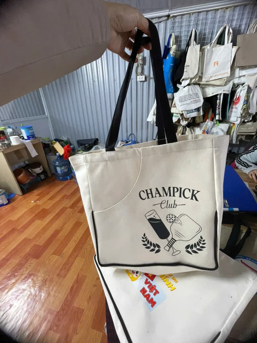 Tư liệu cầm hai quai đen của túi canvas kem với hình CHAMPICK Club ở mặt trước