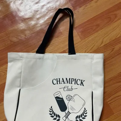 Ảnh thật mặt trước túi canvas kem, hai quai đen, hình CHAMPICK Club và viền đen chữ U