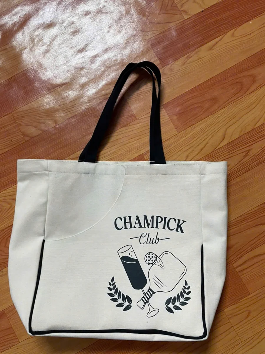Ảnh thật mặt trước túi canvas kem, hai quai đen, hình CHAMPICK Club và viền đen chữ U